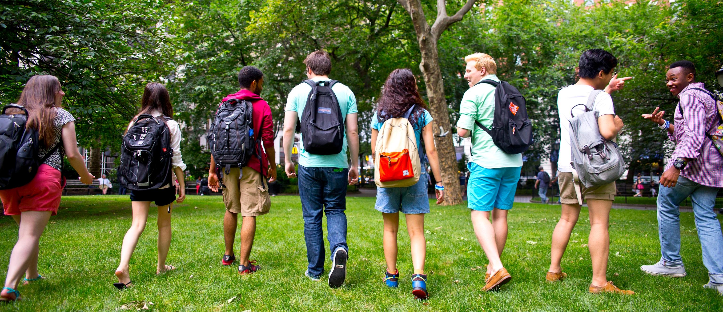 A group of eight people walking across a grassy tree-lined area with their backs to the camera, talking together, all wearing backpacks of various styles and colors